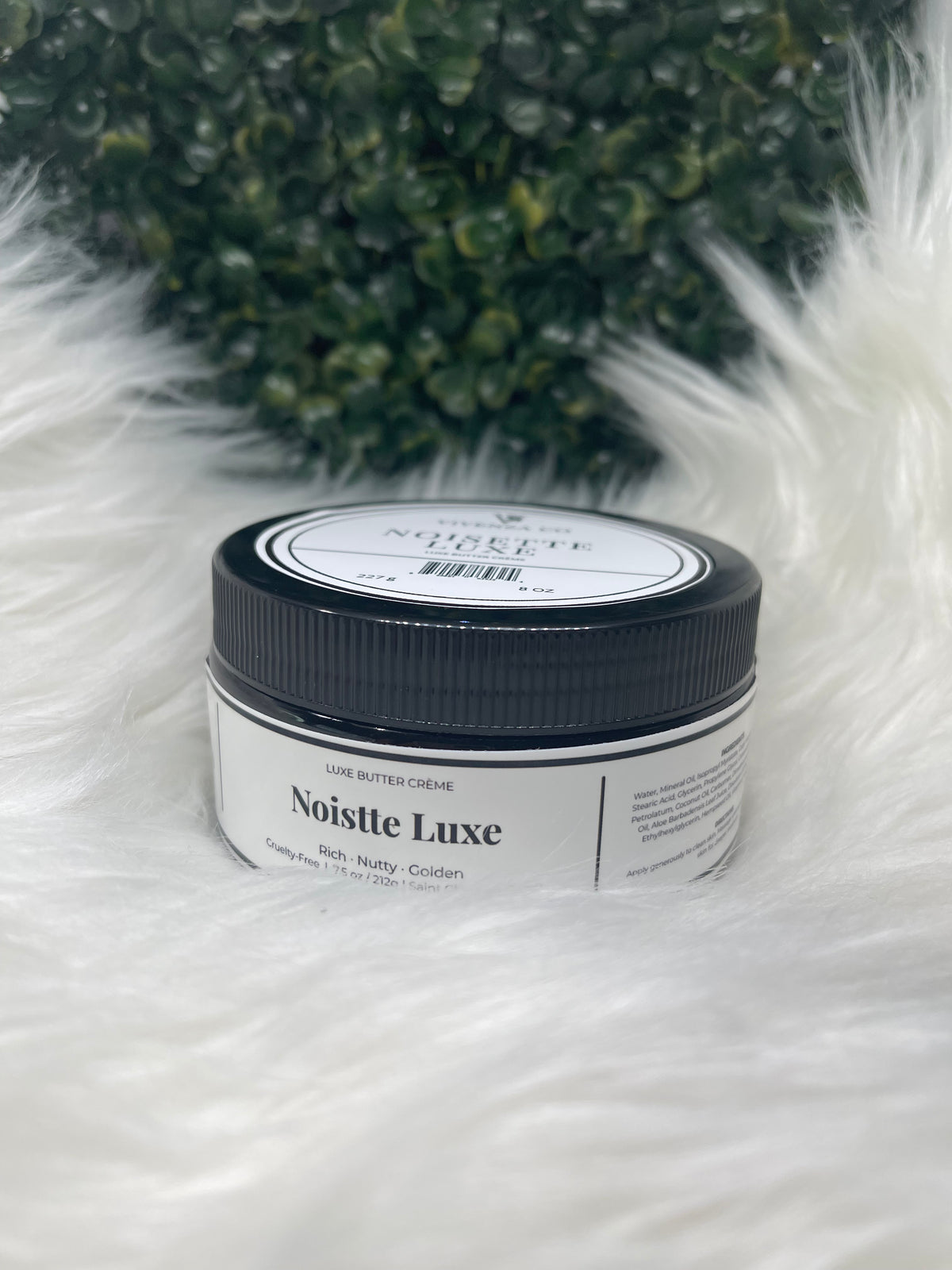 Jar of 'Noisette Luxe' cream on a fluffy white surface with green decorative elements.