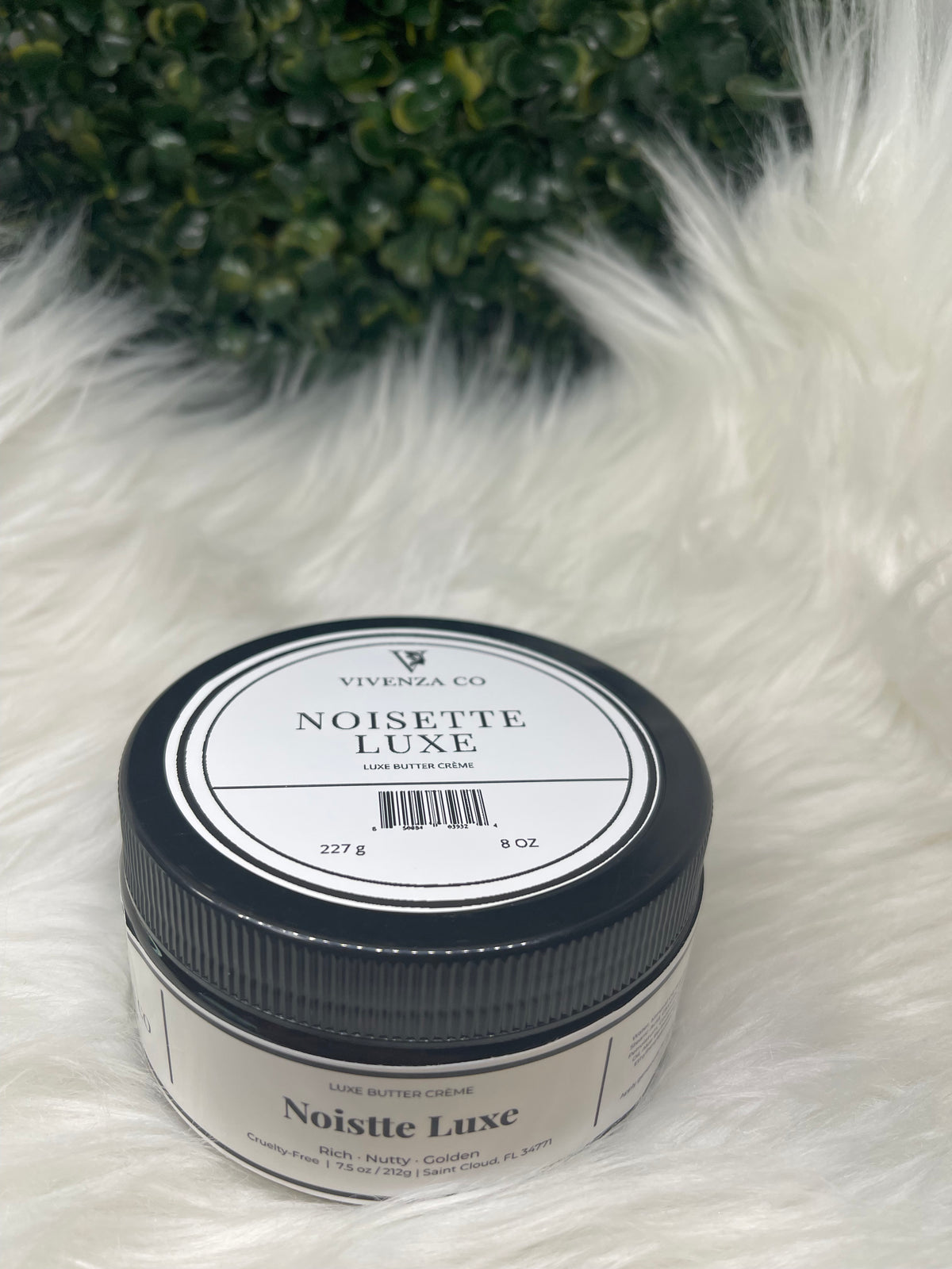 Jar of Noisette Luxe on a fluffy white surface with greenery in the corner