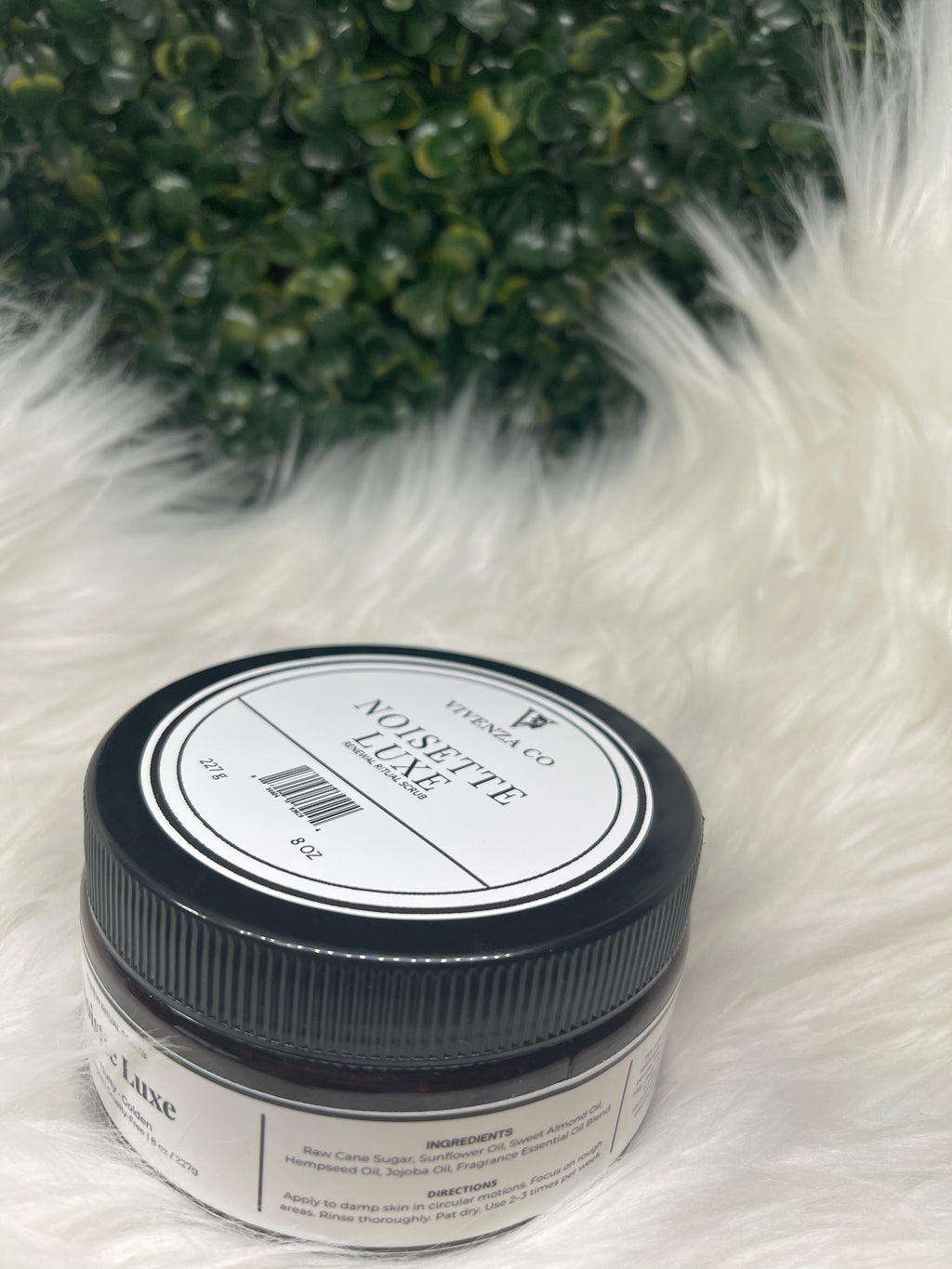 Jar of 'Noisette Luxe' on a fluffy white surface with greenery in the corner