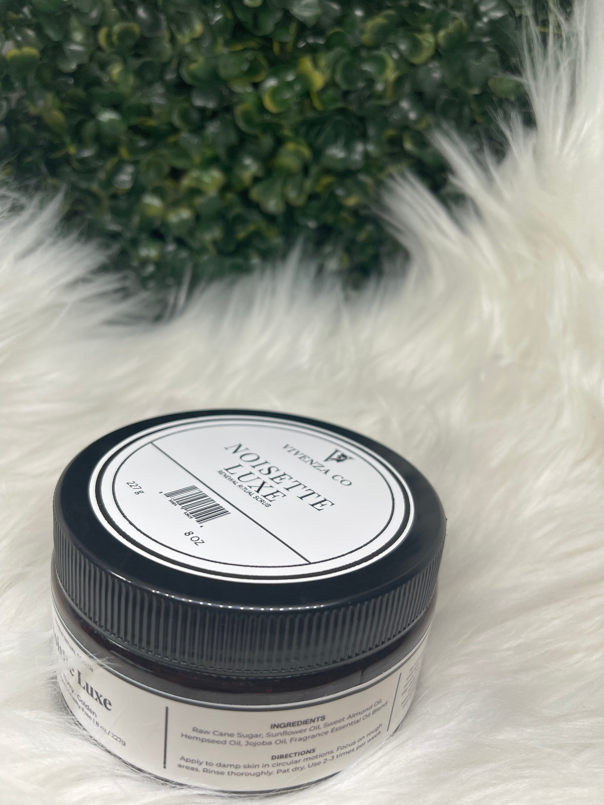 Jar of 'Noisette Luxe' on a fluffy white surface with greenery in the corner