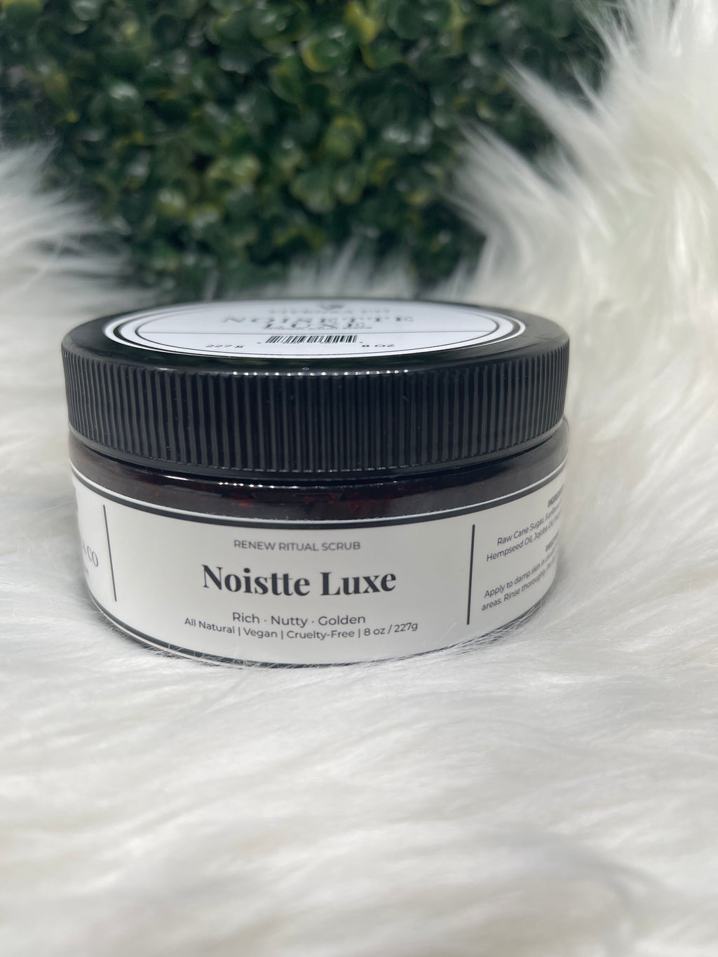 Jar of Noisette Luxe on a fluffy white surface with greenery in the background