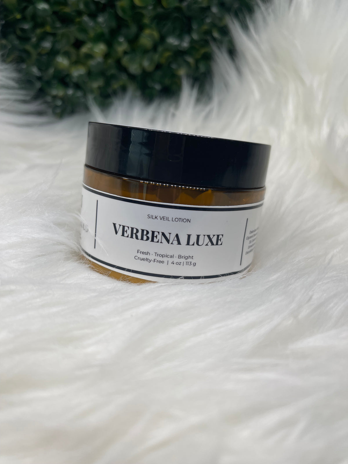 Jar of Verbena Luxe cream on a fluffy white surface with greenery in the corner