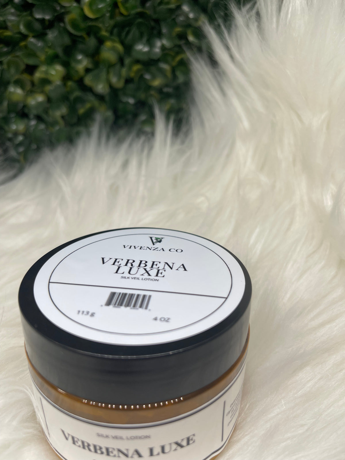 Jar of Verbena Luxe product on a white fur surface with green leaves in the foreground