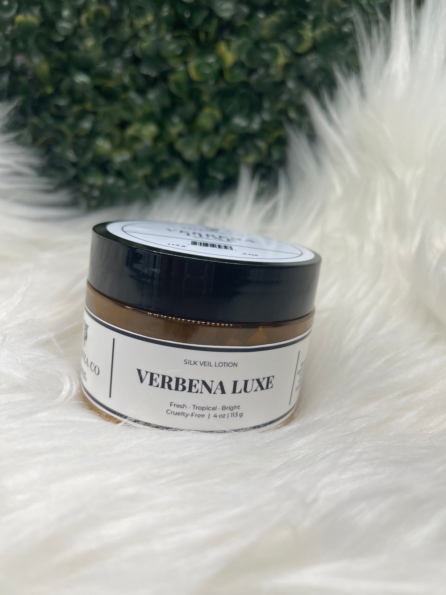 Jar of Verbena Luxe product on a fluffy white surface with green leaves in the corner. 