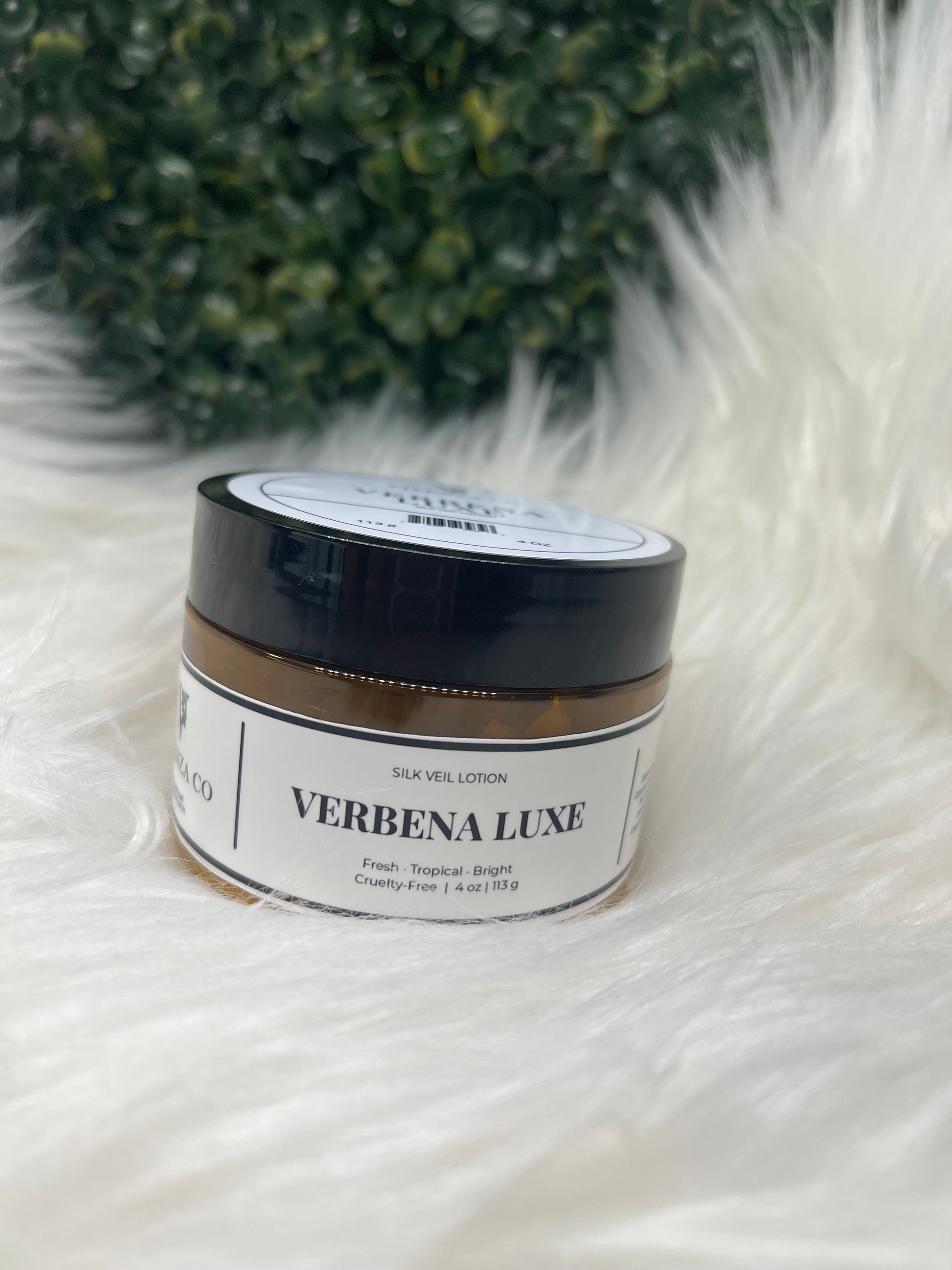 Jar of Verbena Luxe product on a fluffy white surface with green leaves in the corner. 
