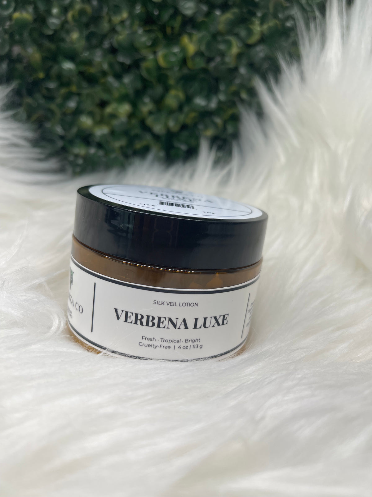Jar of Verbena Luxe product on a fluffy white surface with green leaves in the corner. 